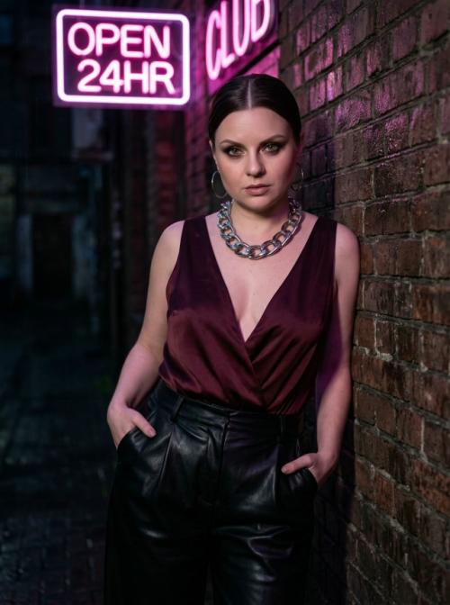 Молодая женщина с идеальной матовой кожей стоит в темном переулке перед неоновой вывеской розового цвета.  